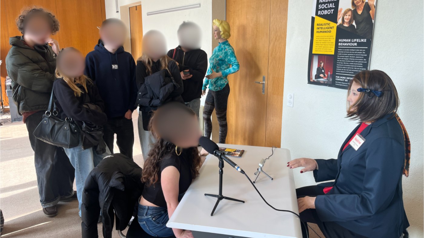 A student sits across the table from Nadine and talks with her while other students watch, faces blurred